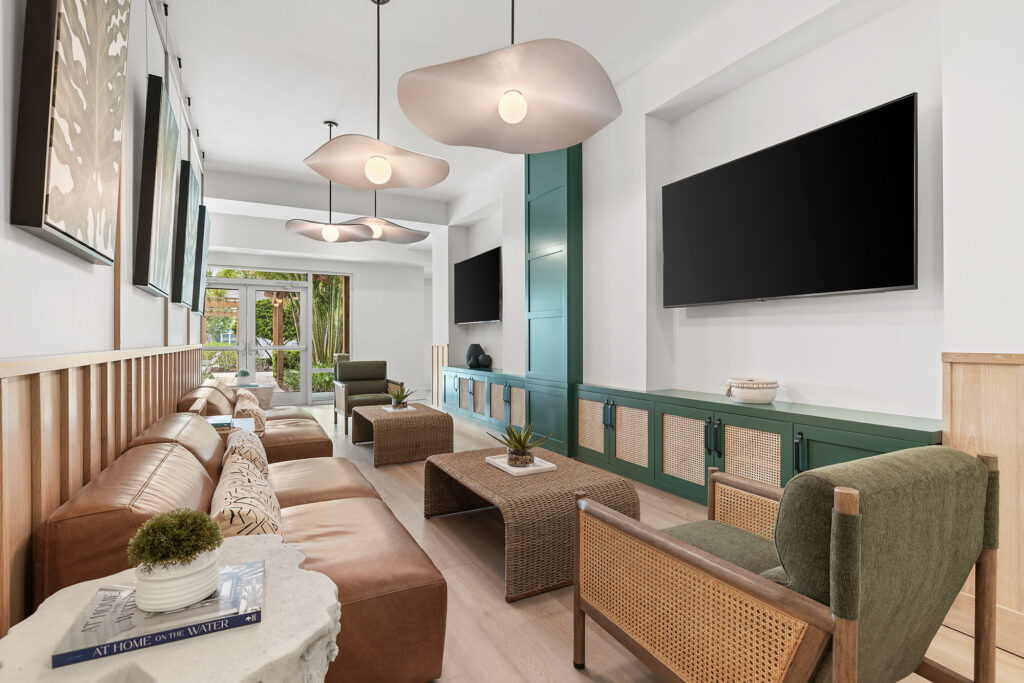 Modern lounge with tan leather sofas, green chairs, two large wall-mounted TVs, unique pendant lights, and a coffee table. Large windows at the back let in natural light; decor includes books and small plants.