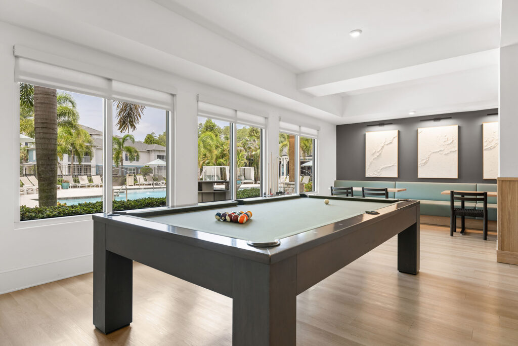 A modern game room with a pool table, cues, and balls, large windows overlooking a pool area with palm trees, and a dining nook with green benches and wall art.