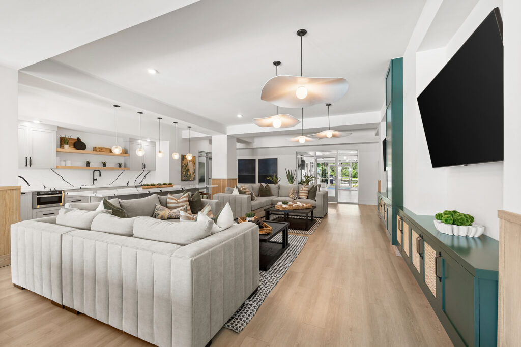 Modern open-concept living room and kitchen with light wood floors, white sofas, pendant lights, a wall-mounted TV, and large windows allowing natural light to fill the space.