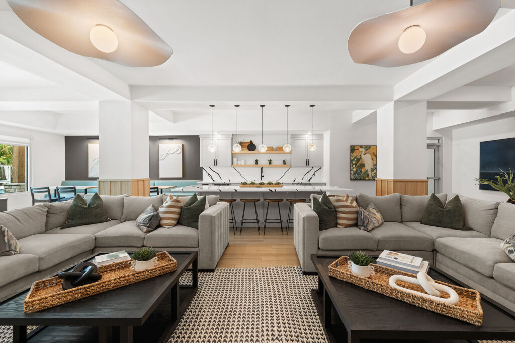 Modern open-concept living room with two gray sectional sofas, patterned rug, black coffee tables, and a white kitchen in the background with pendant lights and barstools. Neutral tones and contemporary décor throughout.