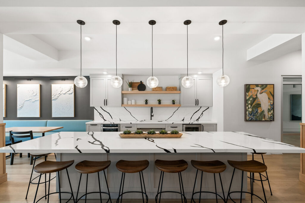 Modern kitchen with a large marble island, six wooden stools, pendant lights, white cabinets, open shelves, abstract wall art, and light wood flooring.