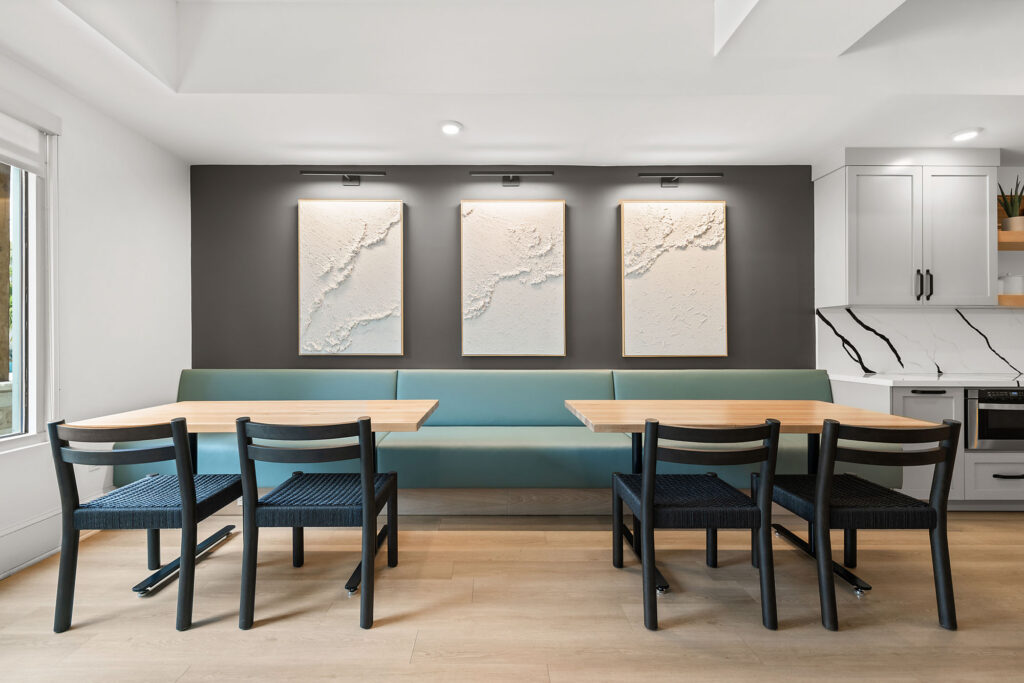 Modern dining nook with a teal cushioned bench, two wooden tables, six black chairs, and three textured white art pieces on a dark accent wall. Minimalist kitchen with white cabinets is visible on the right.