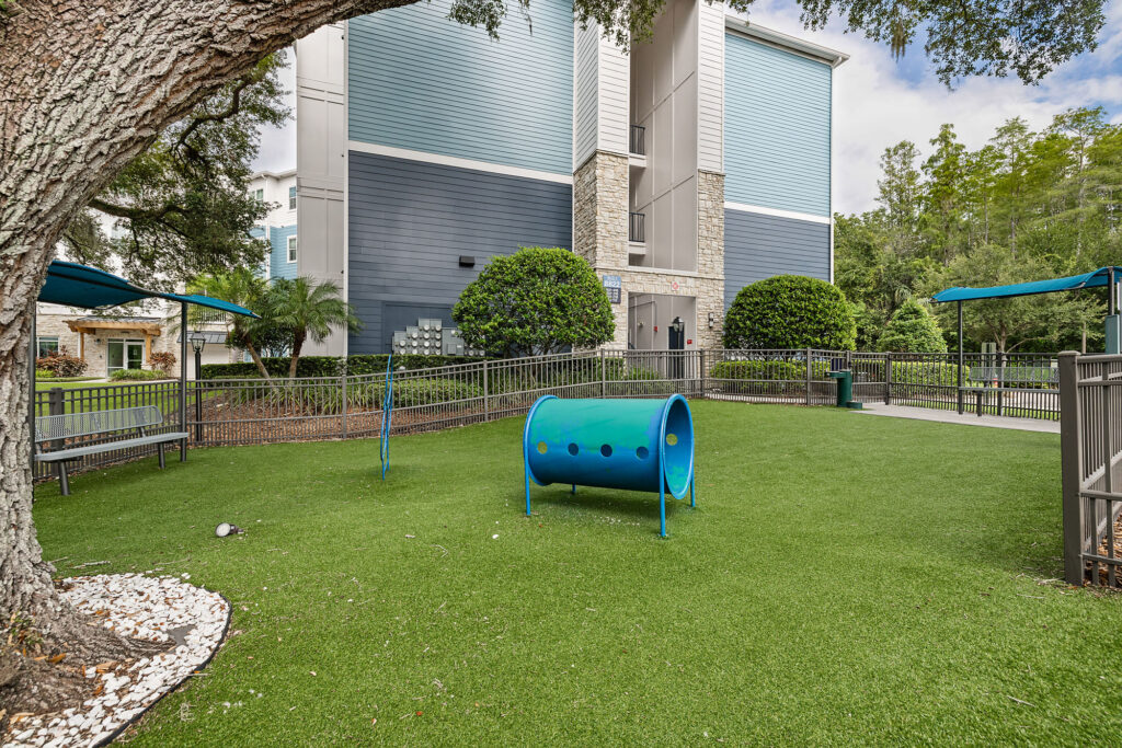 A fenced dog play area with artificial grass, a blue tunnel, shaded benches, and trees. An apartment building is in the background.