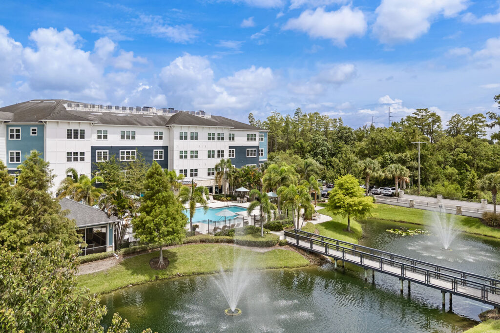 A modern apartment building with a pool, palm trees, and a pond featuring fountains and a footbridge under a partly cloudy sky.