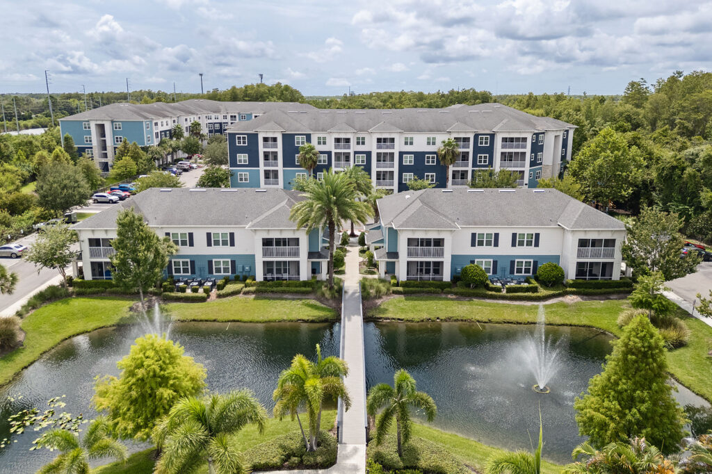 Aerial view of apartment buildings with blue and white exteriors, surrounded by trees, ponds with fountains, and a bridge crossing the water.