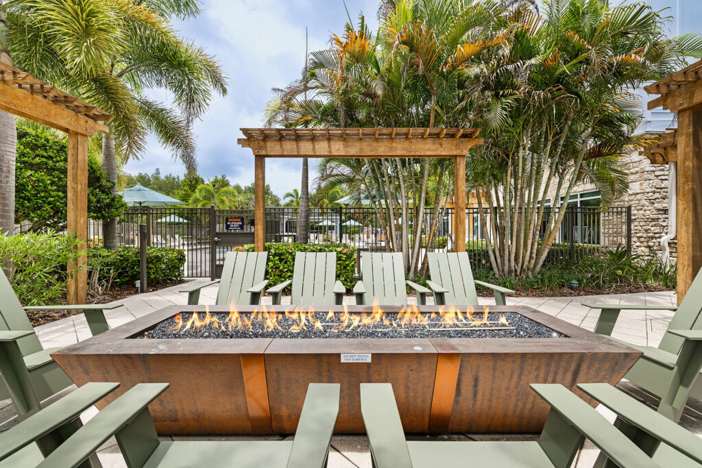 A rectangular fire pit surrounded by green Adirondack chairs and palm trees, set on a tiled patio beneath wooden pergolas, with a gated pool area in the background.