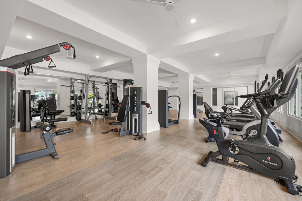 Modern gym with light wood floors, white walls, and large windows. Equipment includes treadmills, stationary bikes, weight machines, and free weights. The space is spacious and well-lit with ceiling fans.