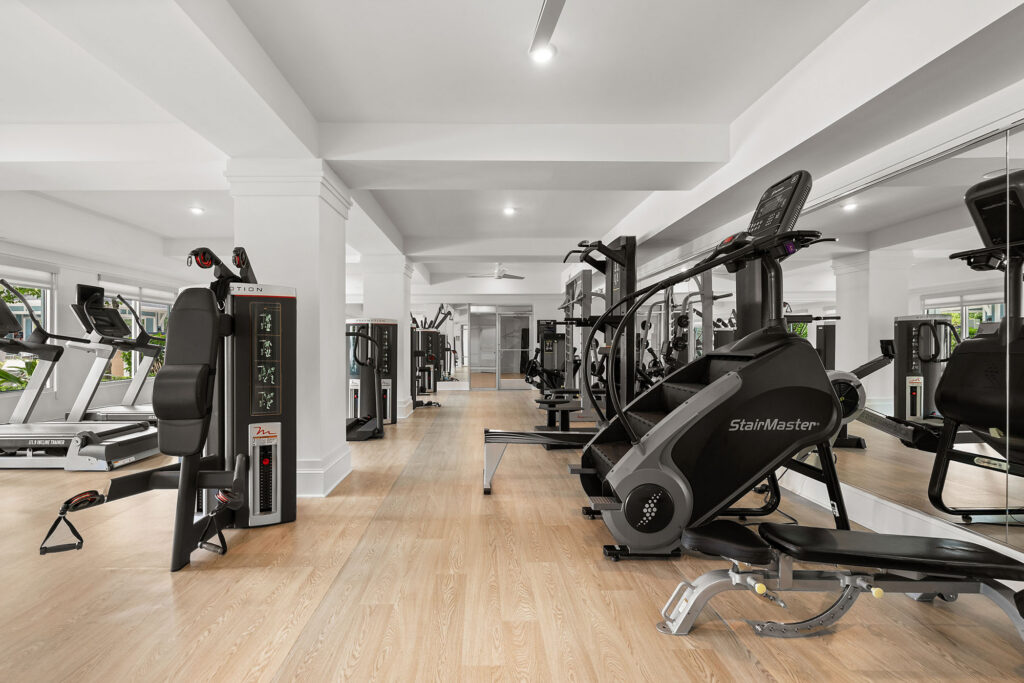 Modern gym with wood flooring, various exercise machines including a StairMaster and weight equipment, large mirrors, white walls, and natural light from multiple windows.