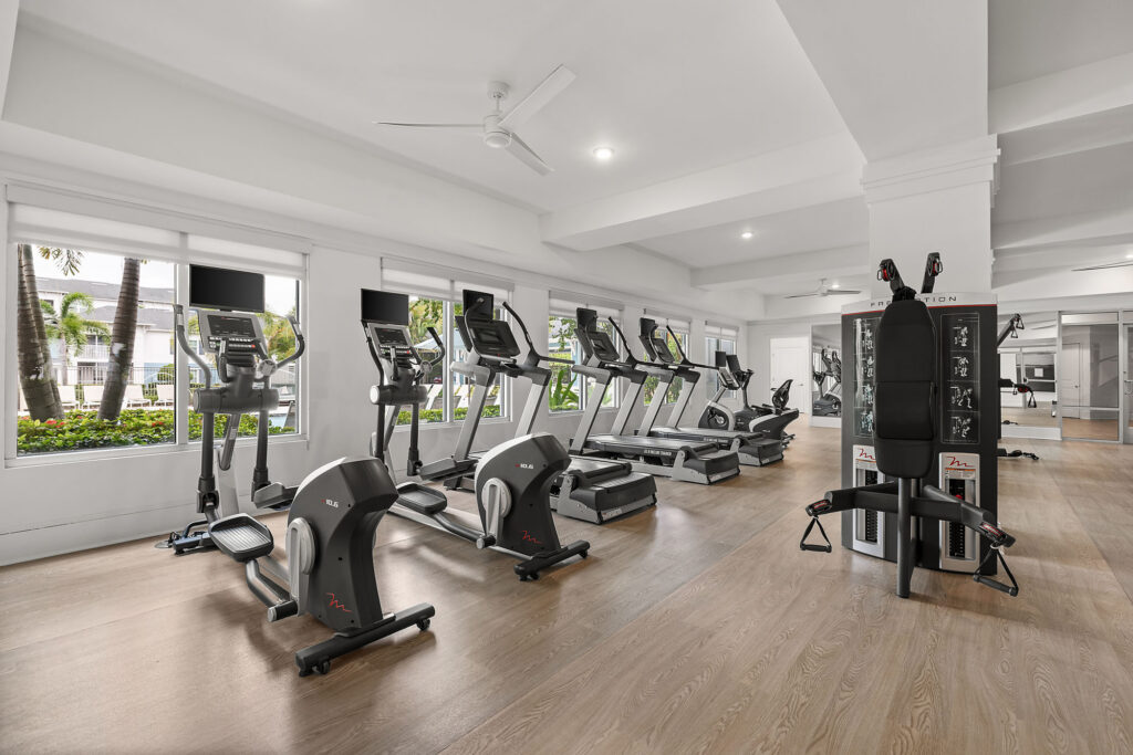 Modern gym with several treadmills, exercise bikes, strength machines, large windows, mirrors, wooden floors, and bright natural light.