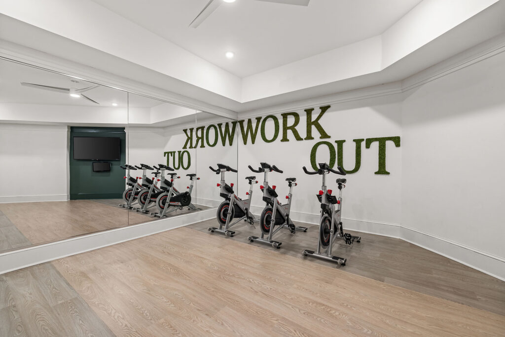 A gym room with four stationary bikes facing a mirror; WORK OUT is written in large green letters on the wall, reflected in the mirror.
