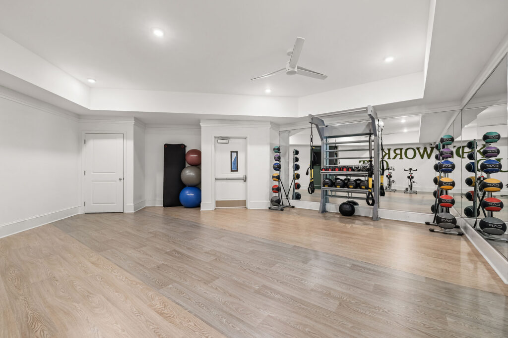 A modern home gym with mirrored walls, exercise balls, a rack of kettlebells, wall-mounted medicine balls, light wood floors, and bright overhead lighting.