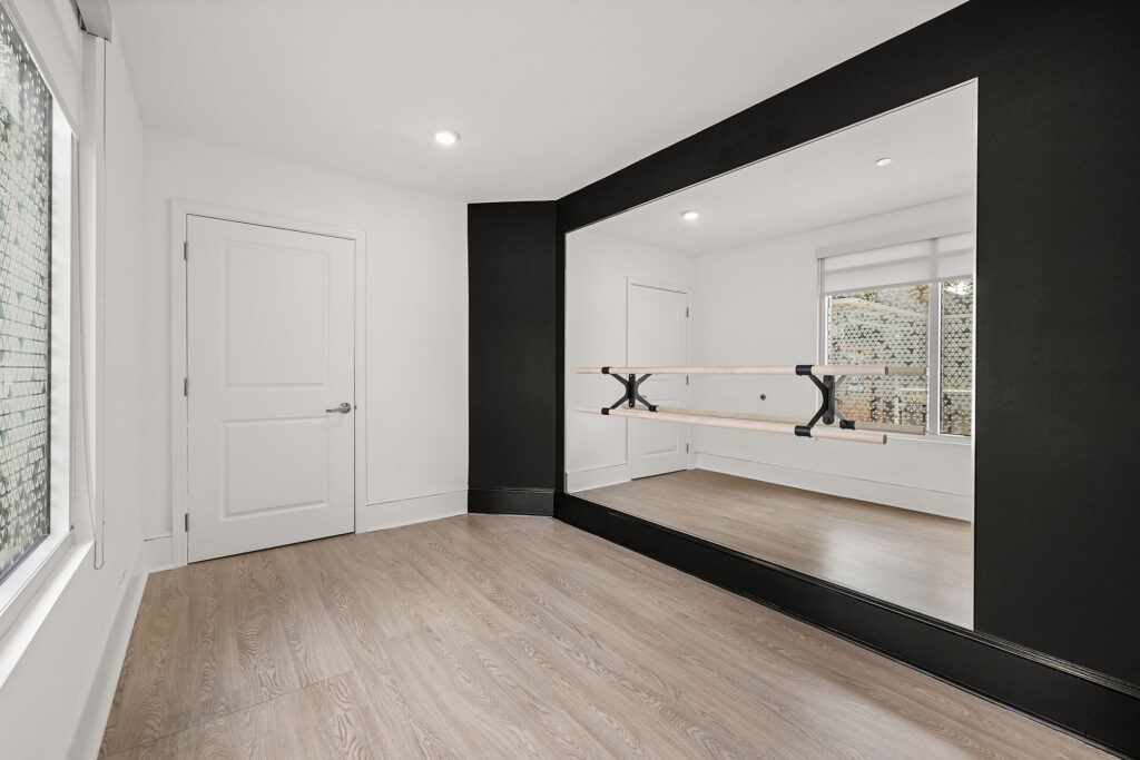A bright, empty room features a large wall mirror with a ballet barre, light wood flooring, white walls, and a window letting in natural light.