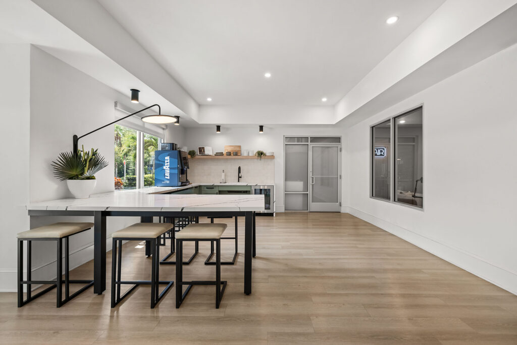 Modern kitchen with white walls, light wood flooring, a table with four stools, wall shelves, and a glass door. Large windows let in natural light, and there are minimalist decor elements.