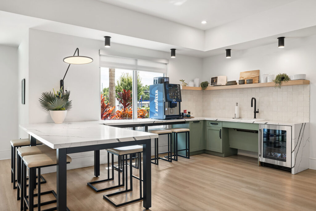 Modern kitchen with white countertops, green cabinets, a coffee machine, barstools, built-in wine cooler, wall shelves, decorative plants, and large windows letting in natural light.