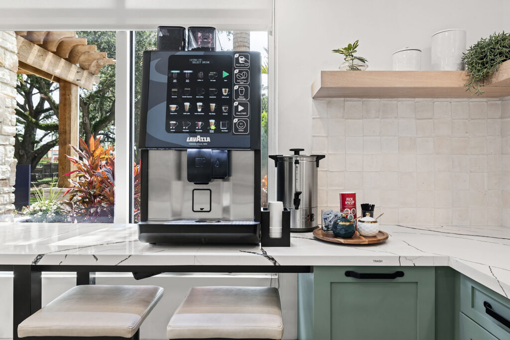 A modern kitchen with a Lavazza coffee machine on a marble counter, coffee supplies on a wooden tray, green cabinets, and a window showing plants outside.