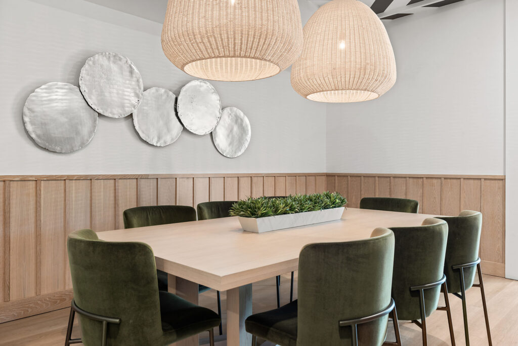 Modern dining room with a light wood table, eight green velvet chairs, woven pendant lights, wood paneling, a rectangular plant centerpiece, and round silver wall art.