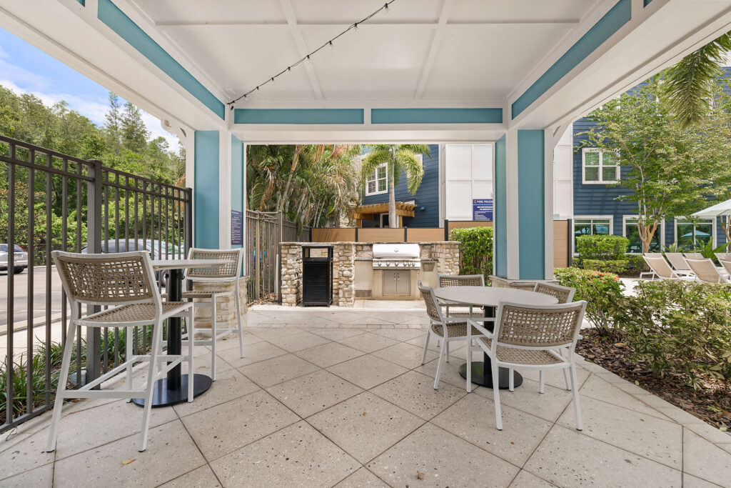 Covered outdoor patio with string lights, two tables and chairs, overlooking a built-in stone grill. Surrounded by greenery, a fence, and blue buildings. Lounge chairs and a pool are visible in the background.