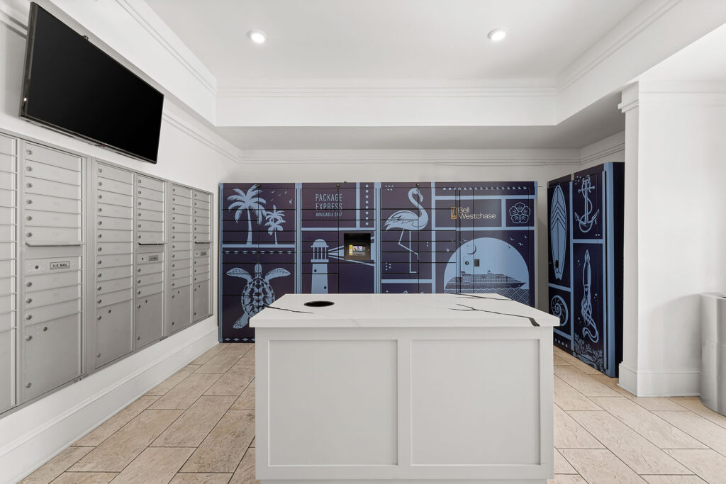 A mailroom with silver mailboxes on the left, a white counter in the center, and blue parcel lockers with travel-themed illustrations on the back wall. A blank TV screen is mounted above the mailboxes.