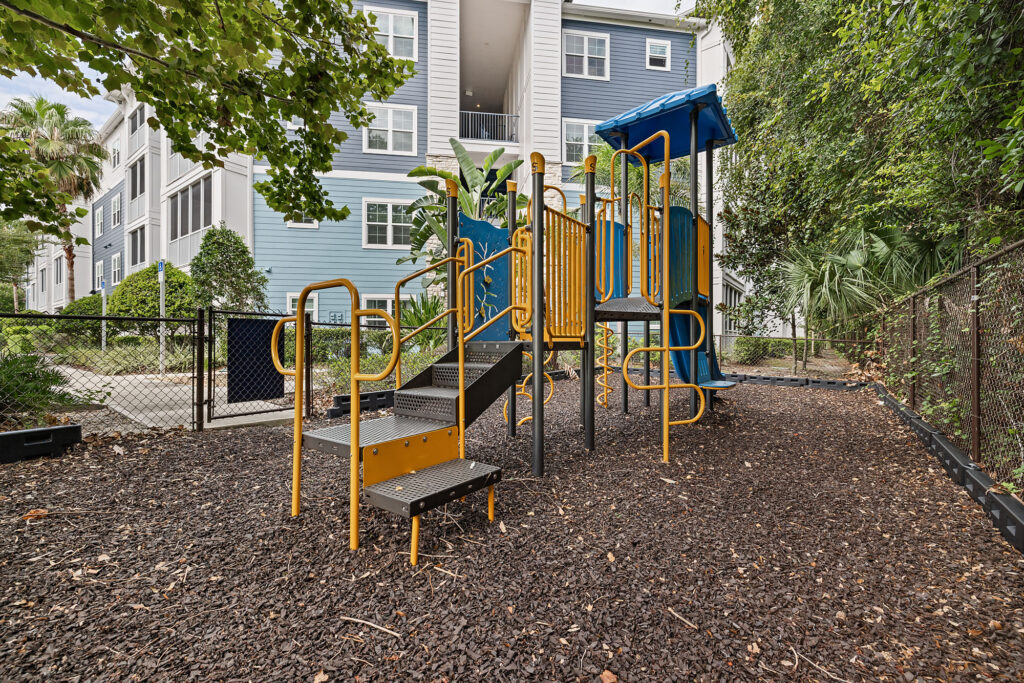 A small playground with yellow and black steps, blue slides, and railings stands on mulch, surrounded by a chain-link fence and greenery, near apartment buildings.