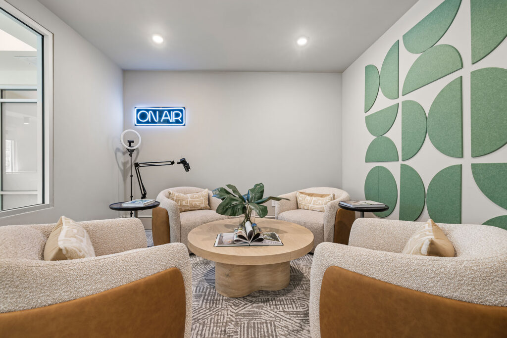 A modern, cozy lounge with four chairs around a round table, green wall art, a plant centerpiece, and a neon ON AIR sign above a tripod light.