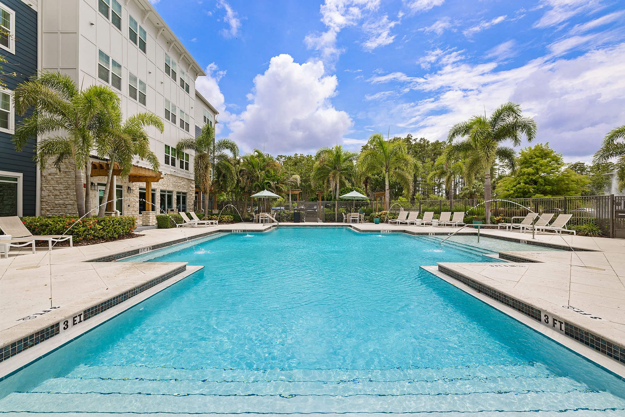 Outdoor swimming pool with clear blue water, surrounded by lounge chairs, palm trees, and a modern apartment building under a partly cloudy sky.
