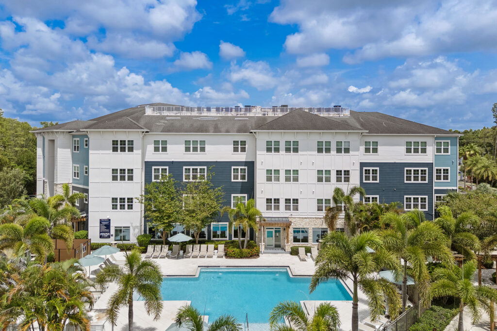 A modern four-story apartment building with blue and white exterior overlooks a large swimming pool, surrounded by palm trees and lounge chairs under a partly cloudy sky.