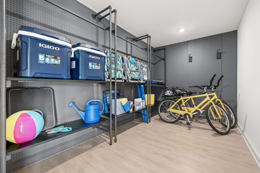 Organized storage room with shelves holding coolers, backpacks, watering cans, and a beach ball. Two yellow bicycles and helmets are next to the shelves on a light wood floor and gray pegboard walls.