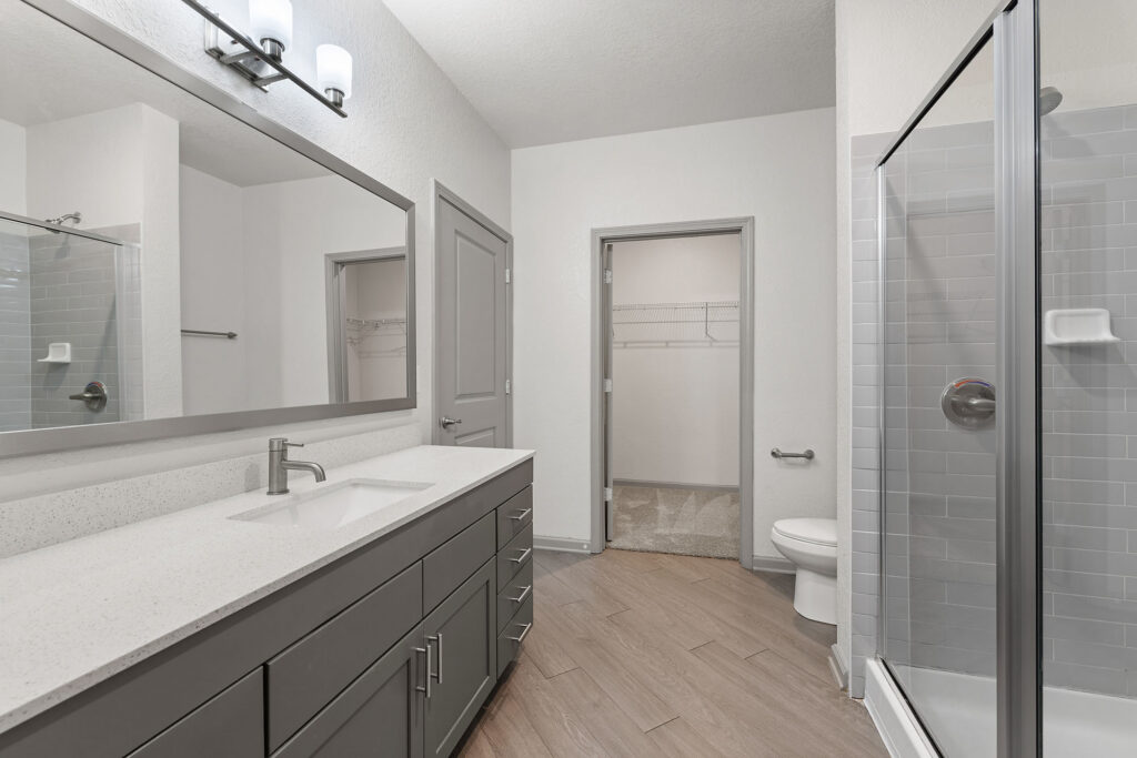 Modern bathroom with gray cabinets, a large mirror, single sink, glass-enclosed shower, white walls, wood-look tile floor, and an open closet seen through a doorway behind the toilet.