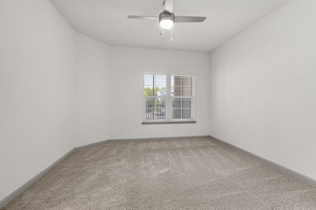 Empty, carpeted room with white walls, a ceiling fan with a light, and a window with blinds letting in daylight.