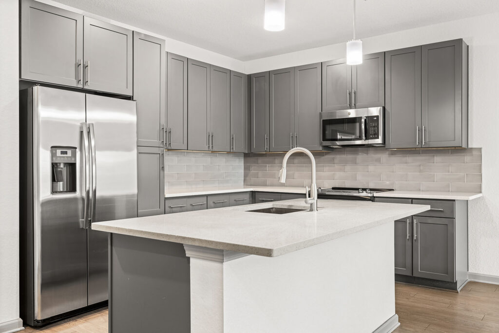 Modern kitchen with gray cabinets, stainless steel appliances, a large island with a sink, subway tile backsplash, and pendant lighting. The space is clean and bright with a neutral color palette.