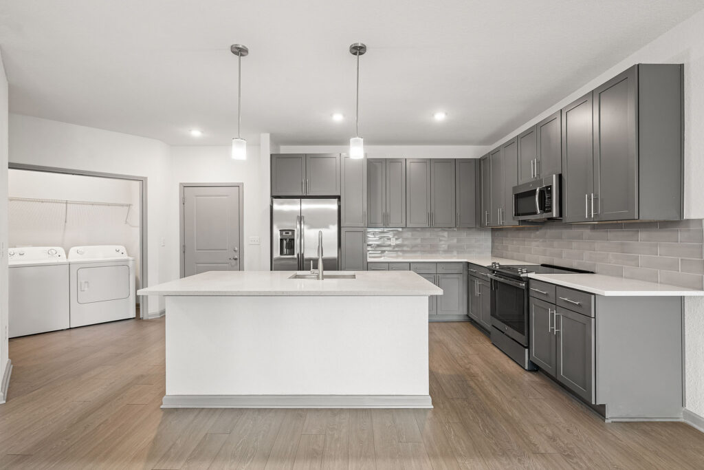 Modern kitchen with gray cabinets, stainless steel appliances, an island with pendant lights, and light wood flooring; a laundry area with washer and dryer is visible to the left.