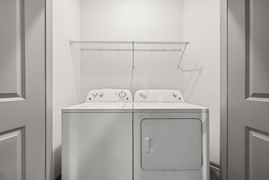 White washing machine and dryer sit side by side in a small laundry closet with white walls and a wire shelf above them. The closet doors are open, revealing the appliances.