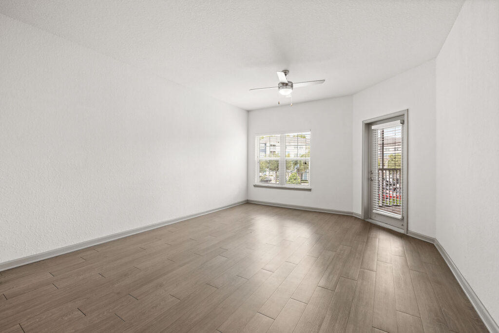 Spacious empty room with light wood flooring, white walls, a ceiling fan, a large window, and a glass door leading to a balcony with greenery visible outside.