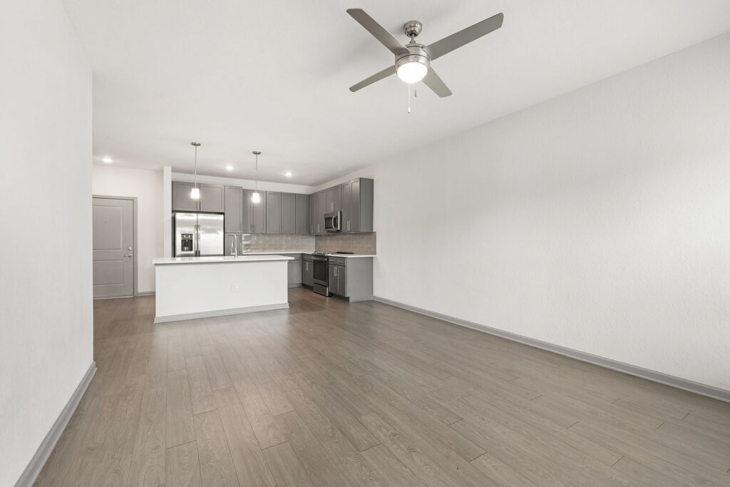 Modern, unfurnished apartment with light gray wood flooring, white walls, kitchen with stainless steel appliances, pendant lights, ceiling fan, and an open floor plan.