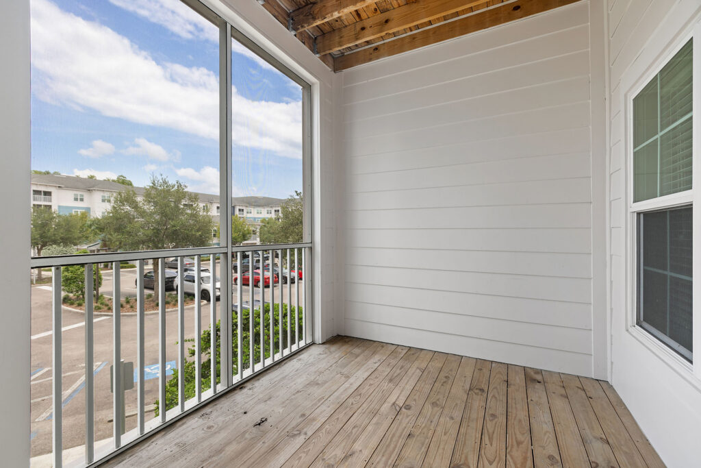 A covered balcony with wooden flooring, white walls, and a railing overlooks a parking lot and apartment buildings under a blue sky with white clouds.